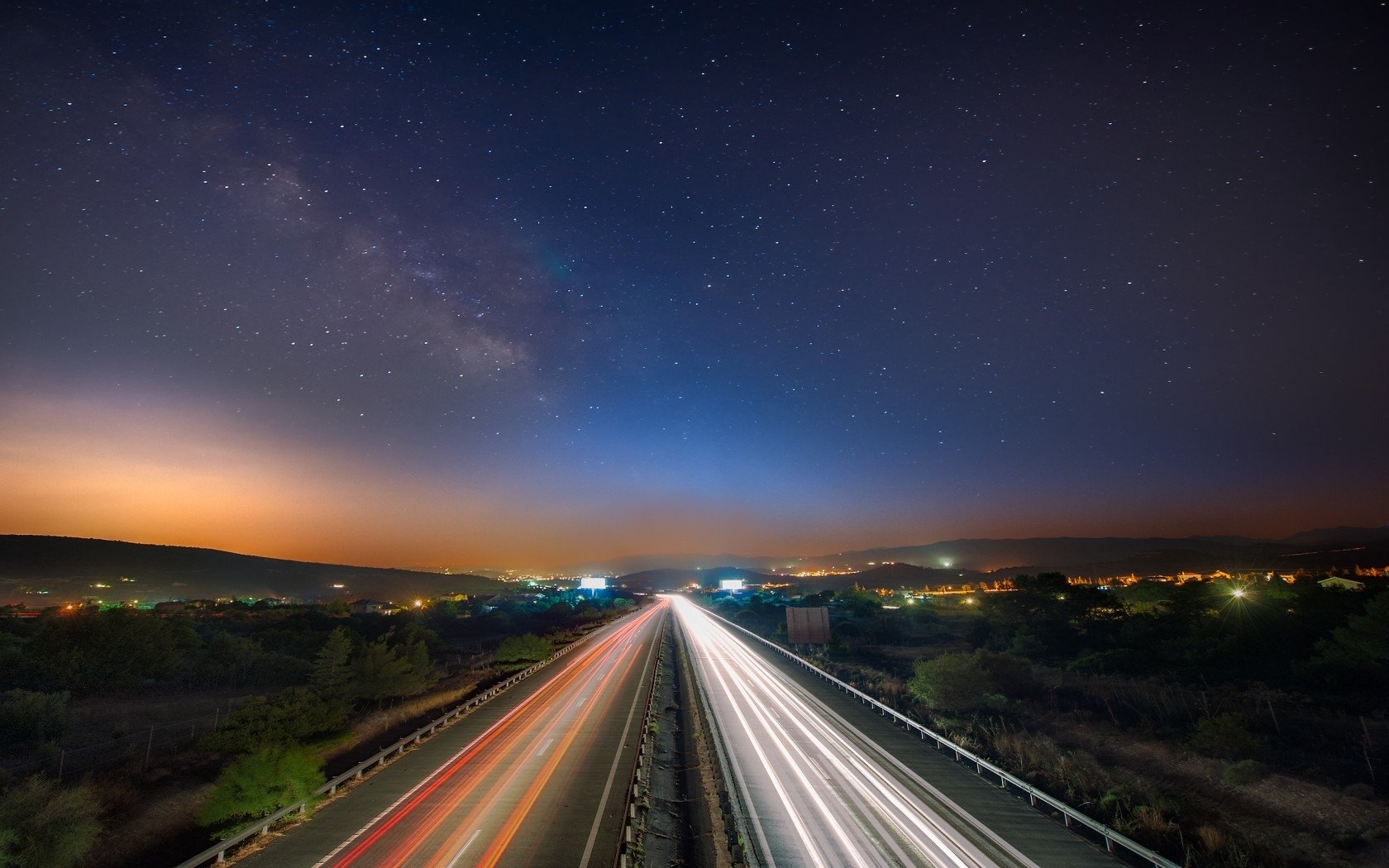 A time lapse photograph capturing the movement of traffic along a highway under a starry sky, highlighting vibrant light trails and a serene landscape at dusk.