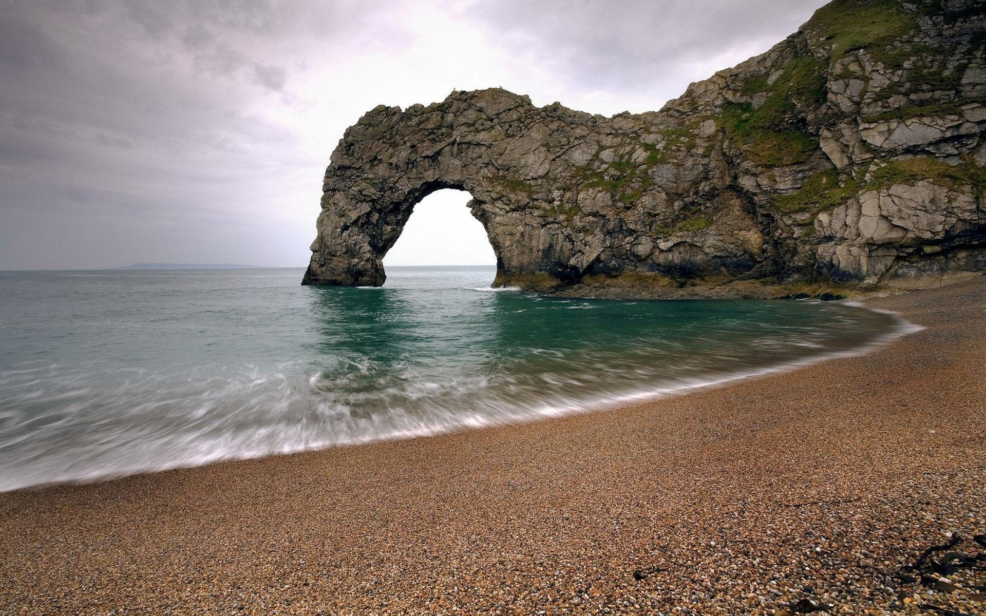 Durdle Door: limestone cliff arch jutting into the sea, gentle waves washing a pebbled beach under a cloudy sky.