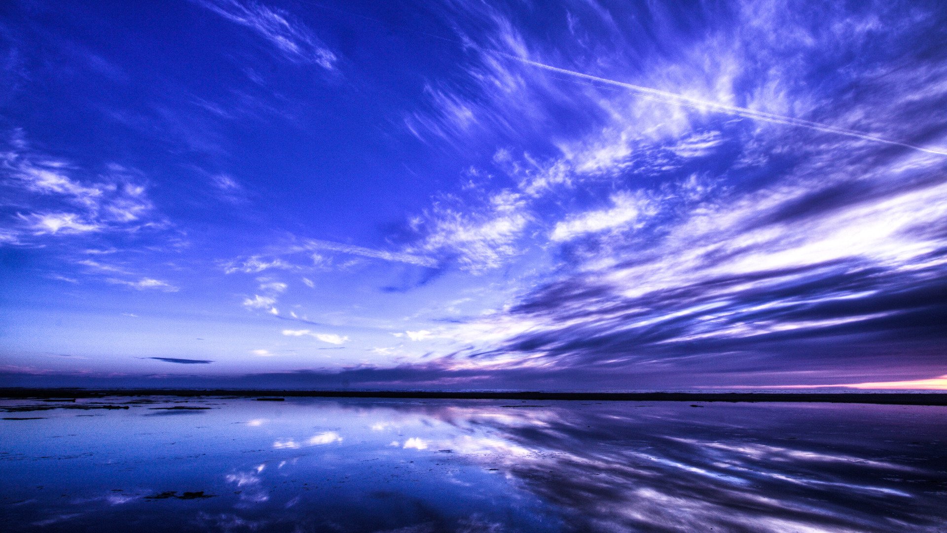 Scenic seascape photograph of a dramatic cloud-filled sky meeting the horizon, its blue-purple hues reflected on calm water.