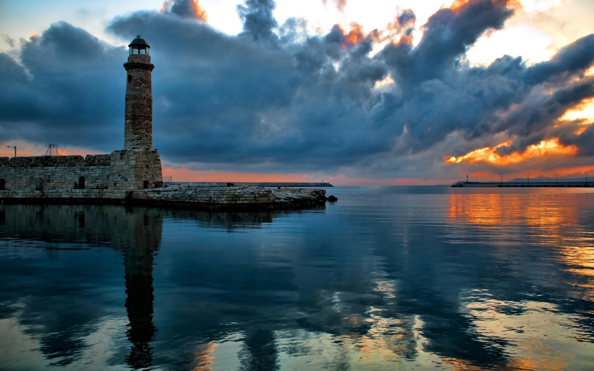 A stunning sunset reflects off calm waters near a man-made lighthouse, silhouetted against dramatic clouds in the sky.