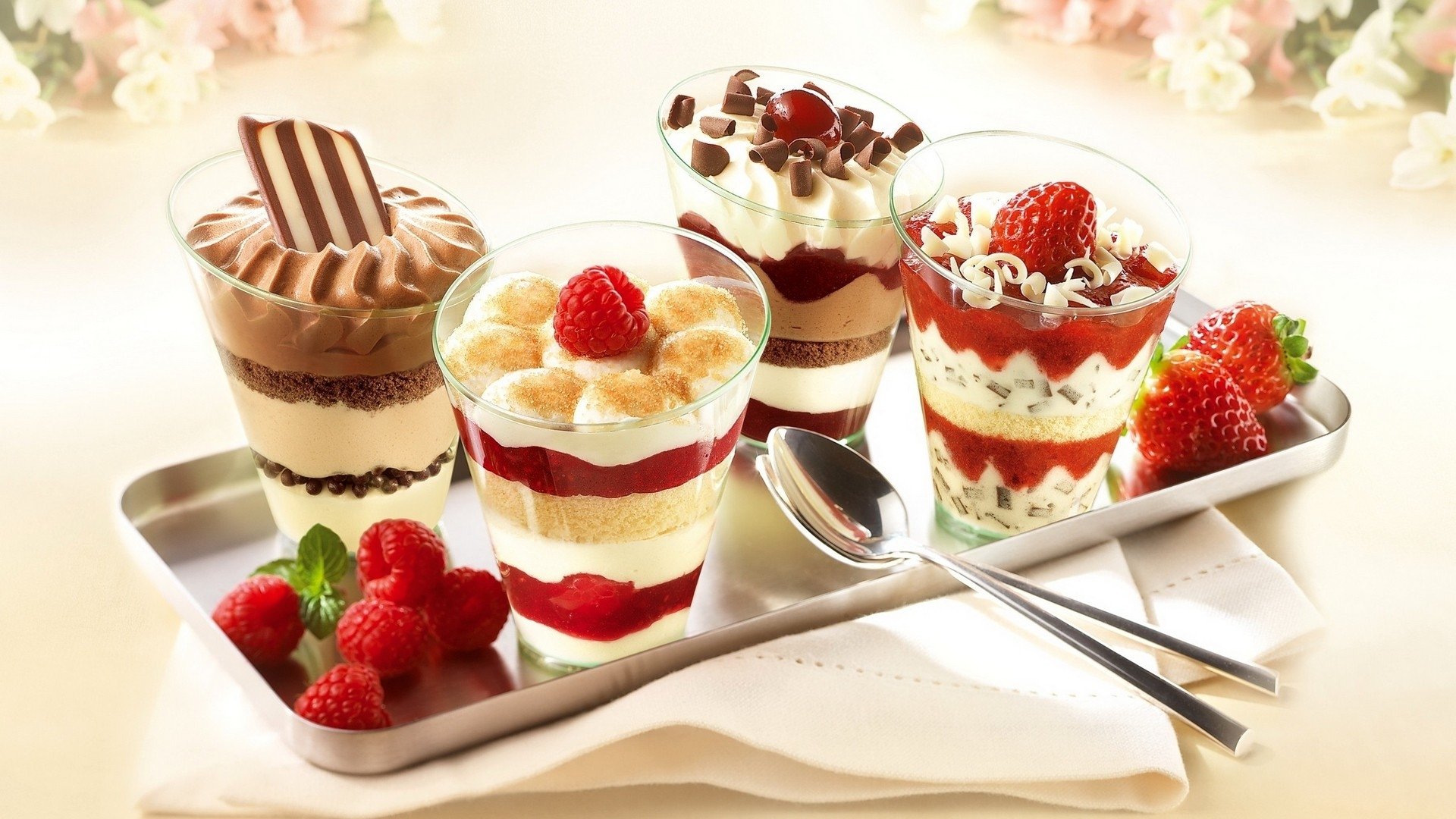 Four beautifully layered desserts in clear cups, featuring fruits, creamy layers, and decorative toppings, served on a silver tray with fresh raspberries beside them.