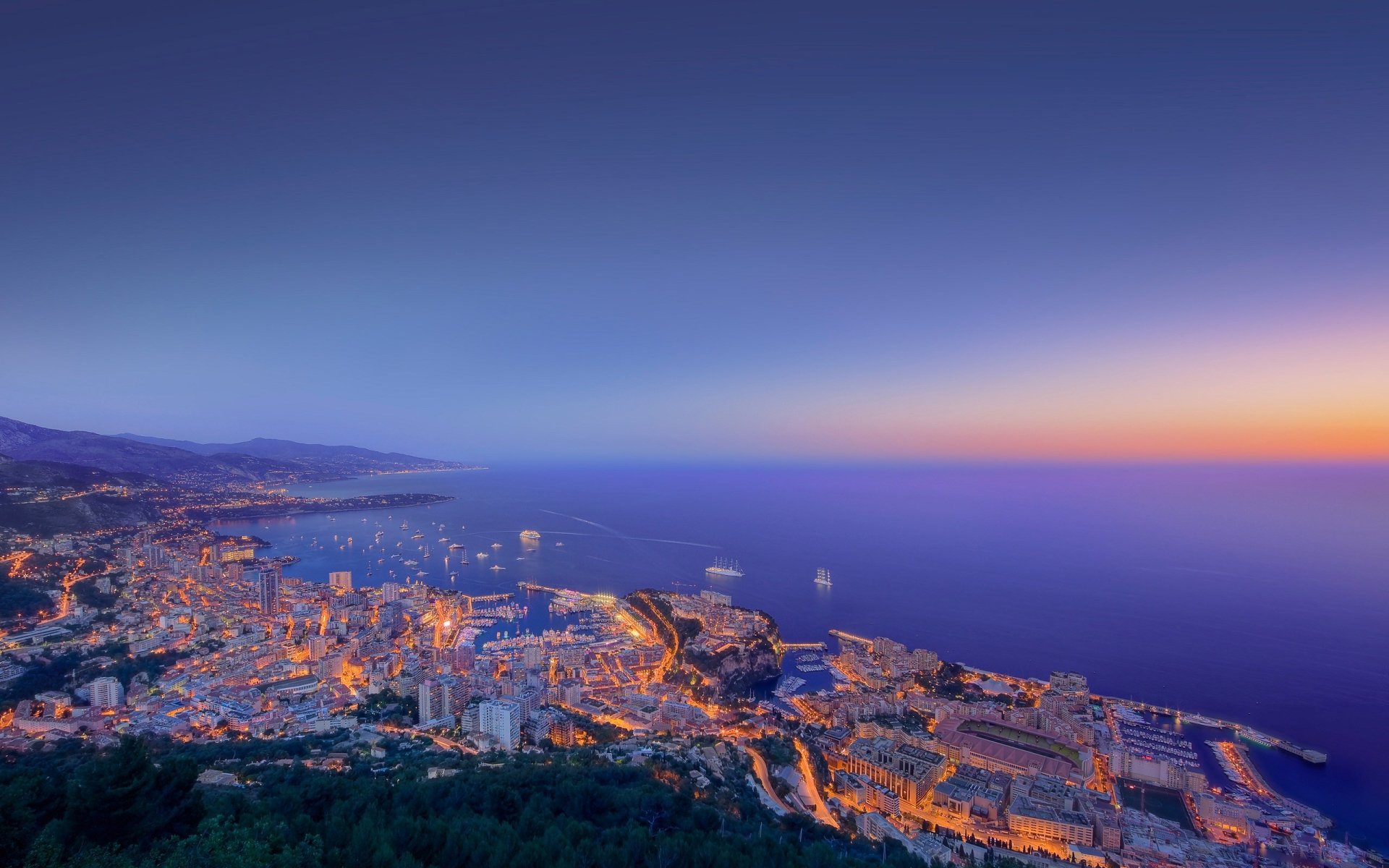 Aerial twilight landscape of Monaco: a man-made coastal cityscape with glowing streets and buildings lining the ocean.
