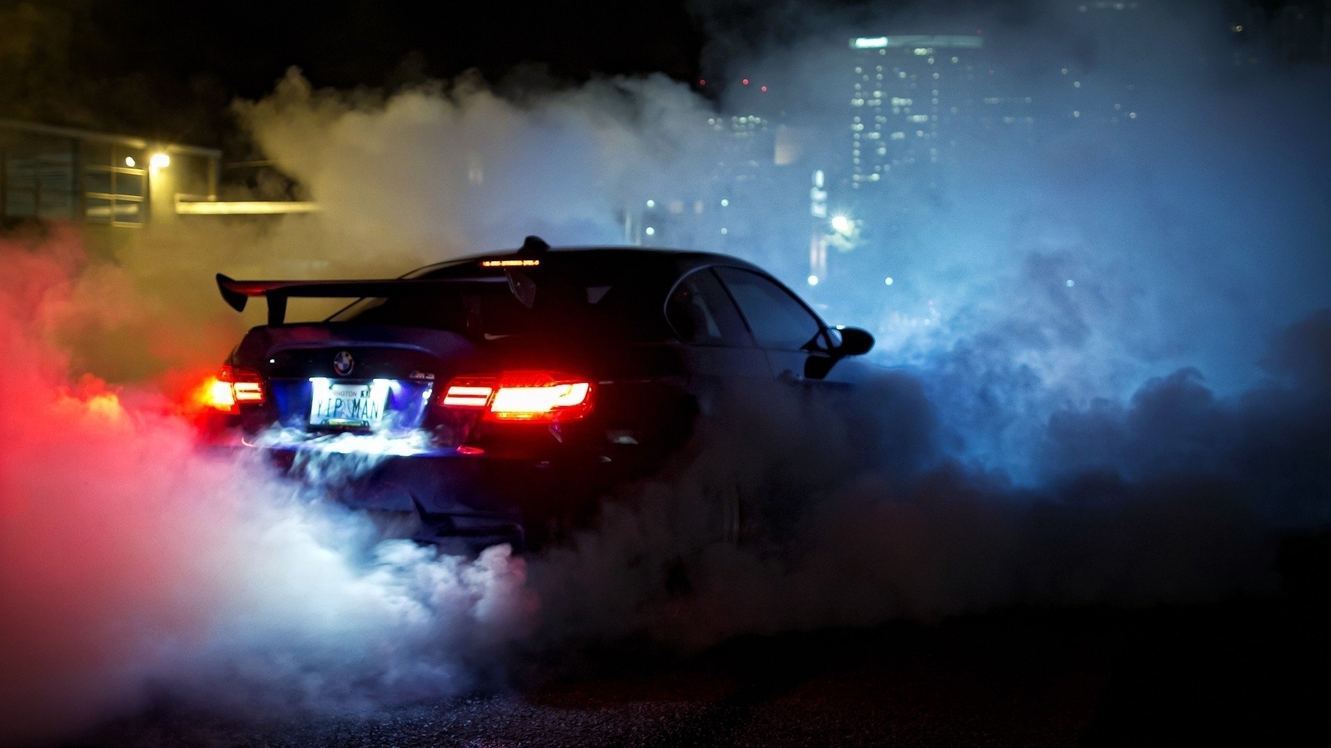 A BMW M3 drifts through a cloud of smoke, illuminated by colorful lights in a nighttime urban setting, showcasing its dynamic performance and sleek design.