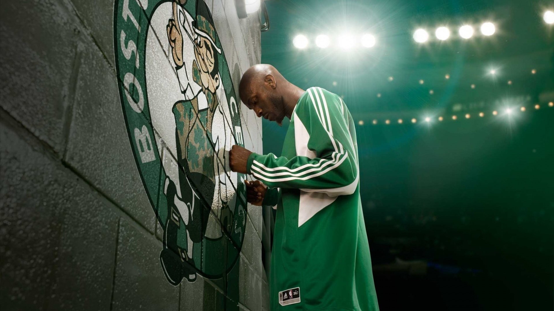 A Boston Celtics player in green warm-up gear stands focused by a large team logo on a wall, illuminated by bright arena lights.