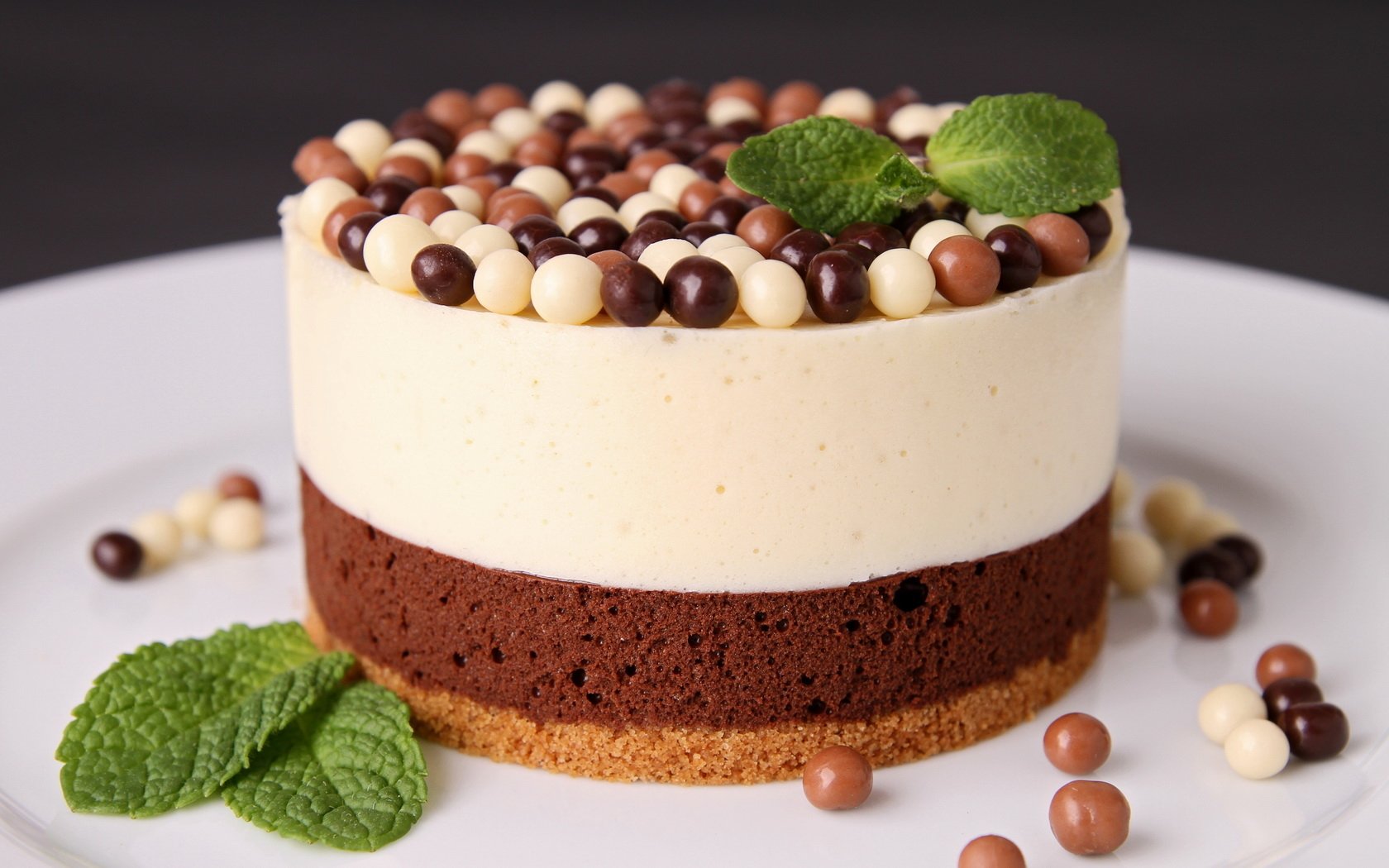 A visually appealing dessert featuring layers of chocolate cake and creamy mousse topped with colorful chocolate balls and fresh mint leaves, served on a white plate.