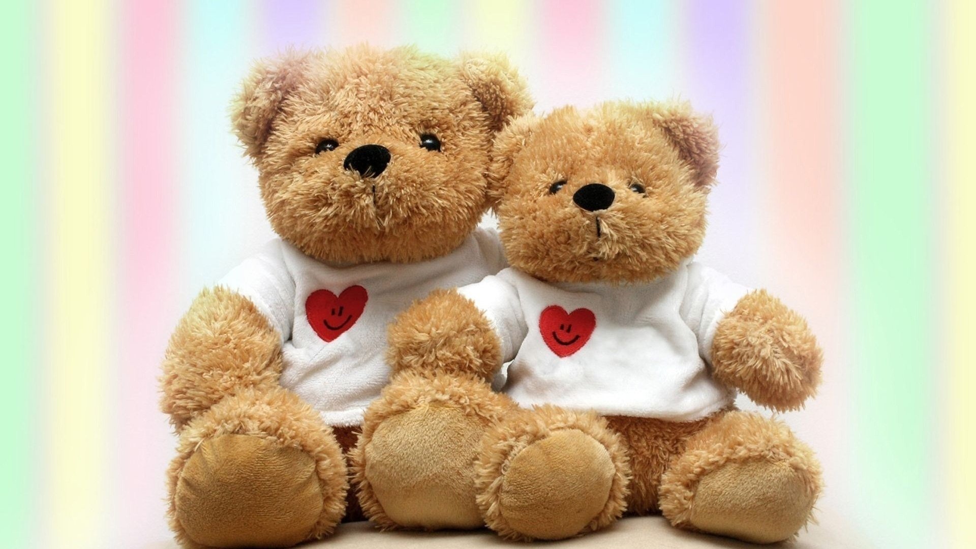 Two brown man-made stuffed animal teddy bears in white shirts with red hearts, sitting side by side against a soft pastel striped background.