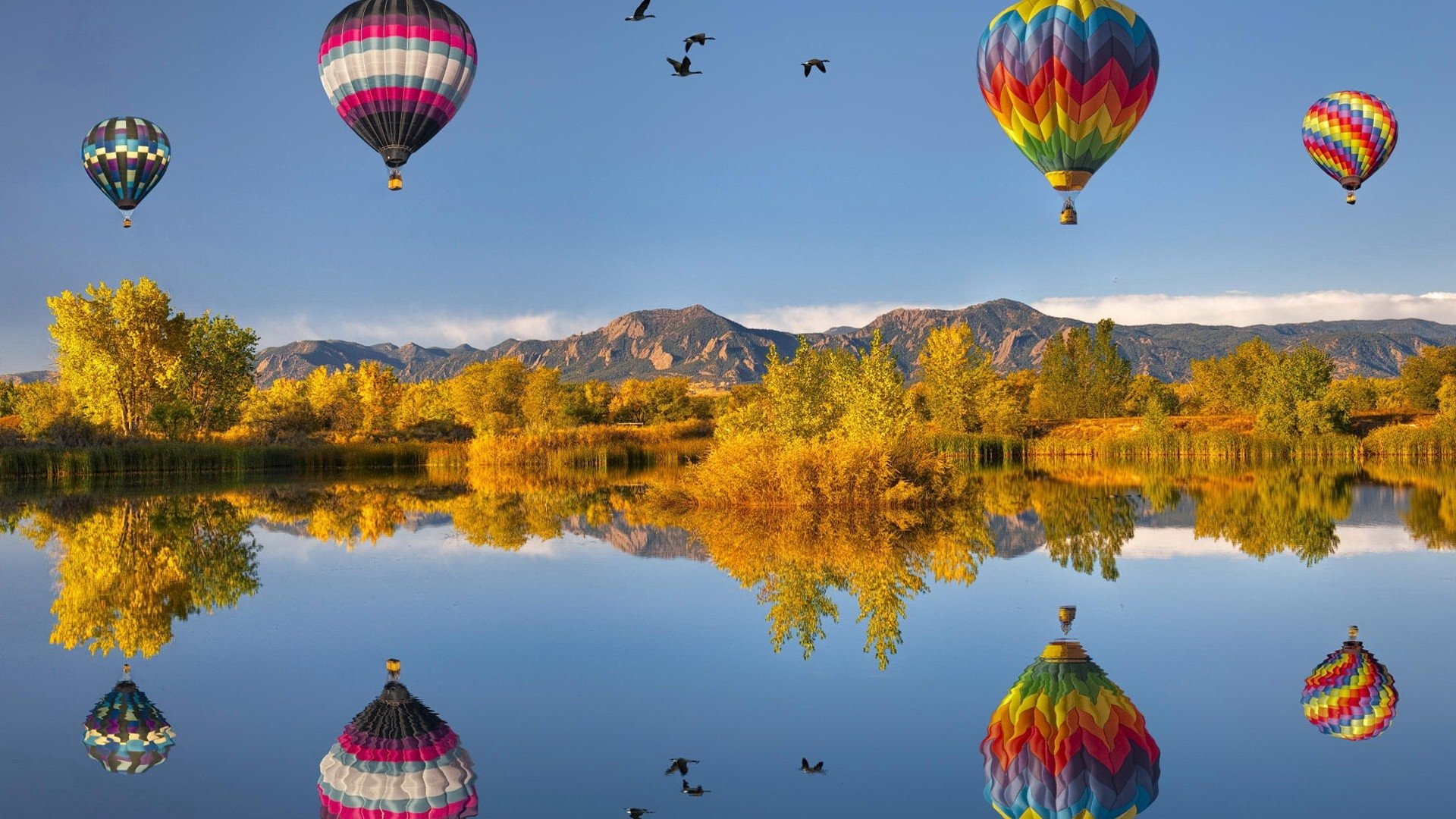 Colorful hot air balloons float above a calm lake, with their reflections mirrored in the water and mountains and trees in the background.