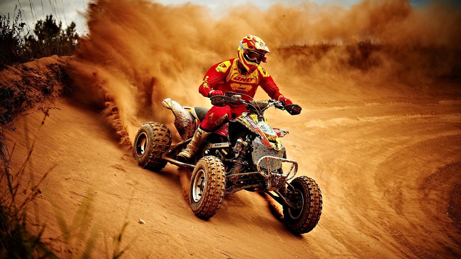 A rider in red gear maneuvers a Can-Am vehicle through a dusty, sandy terrain, kicking up a cloud of dirt behind.