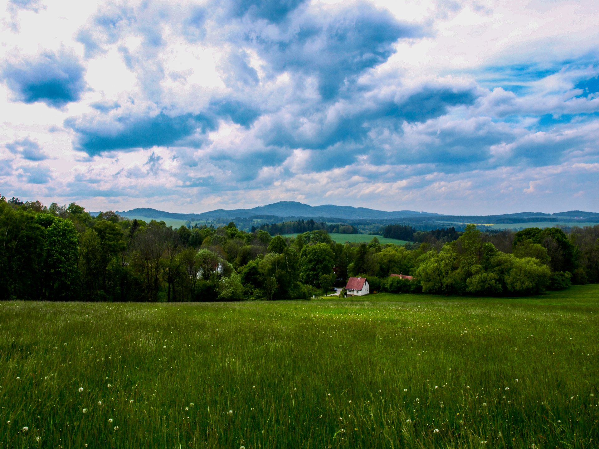 Landscape nature photograph in the Czech Republic: a green meadow stretching to a small house with a red roof among trees, rolling hills under a dramatic cloudy sky.