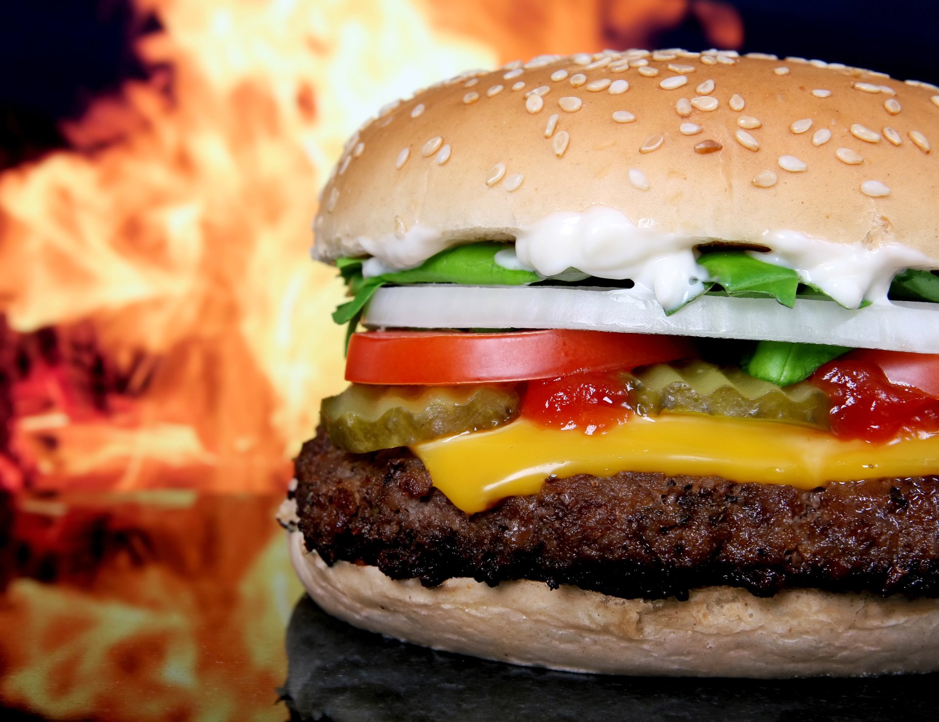 Close-up of a cheeseburger with lettuce, tomato, onion, pickles, and condiments in a sesame seed bun with flames in the background.