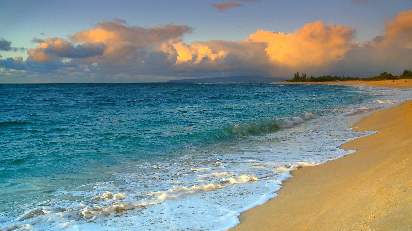 A serene beach in Hawaii with gentle waves lapping at the shore, under a sky filled with soft clouds and warm colors, capturing the beauty of the ocean landscape.