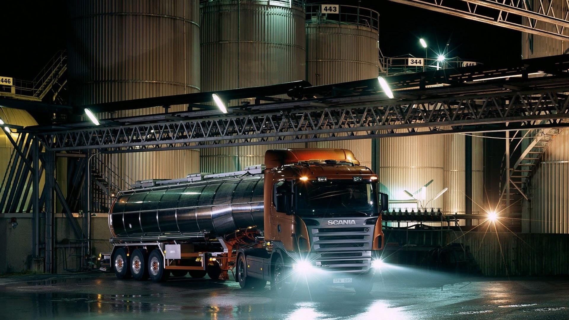A Scania vehicle stands illuminated at night, with bright headlights and a sleek design, parked near large industrial silos and machinery in a manufacturing setting.