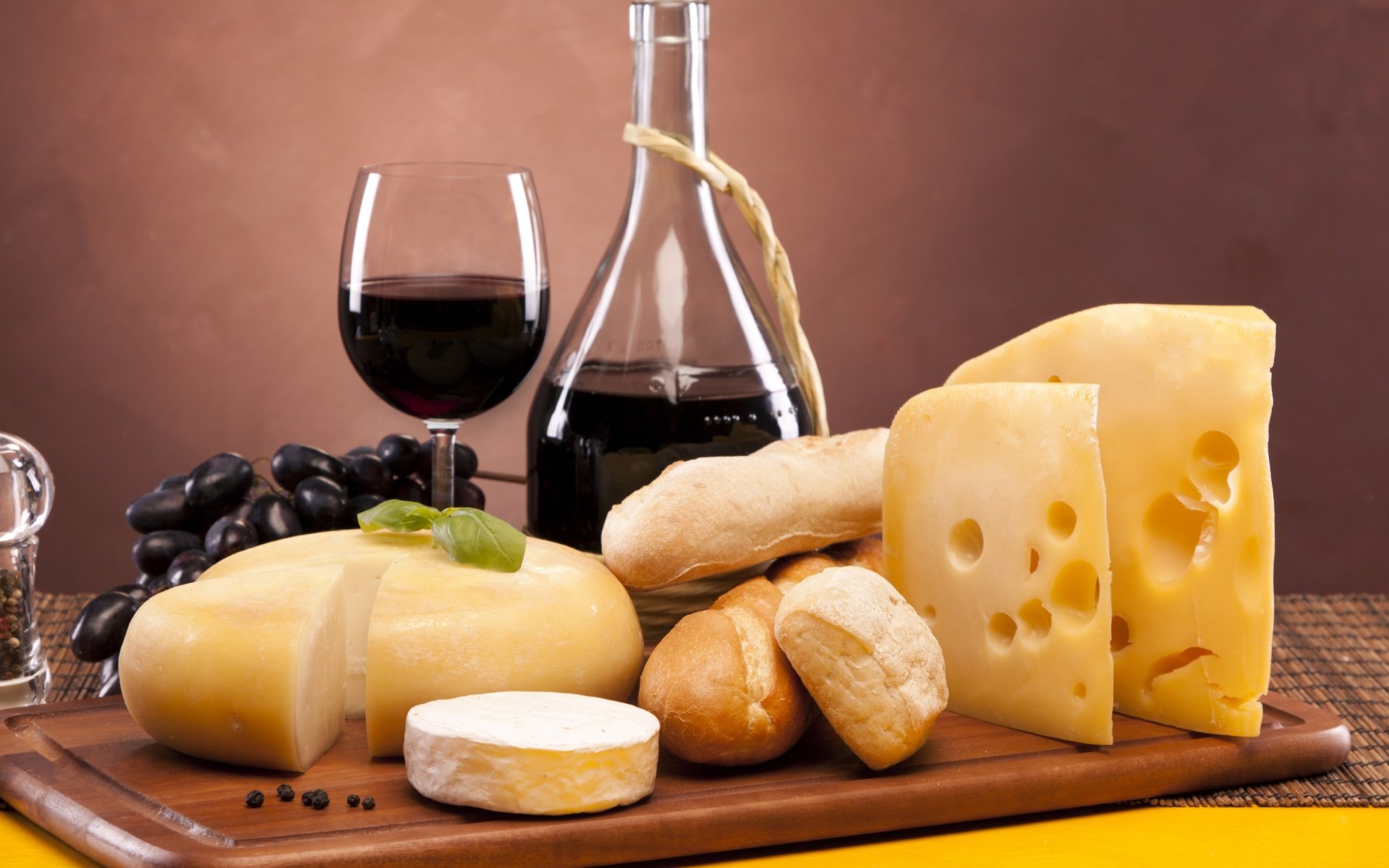 Still life of various cheeses, bread rolls, a glass and decanter of red wine, and black grapes arranged on a wooden board against a warm brown background.