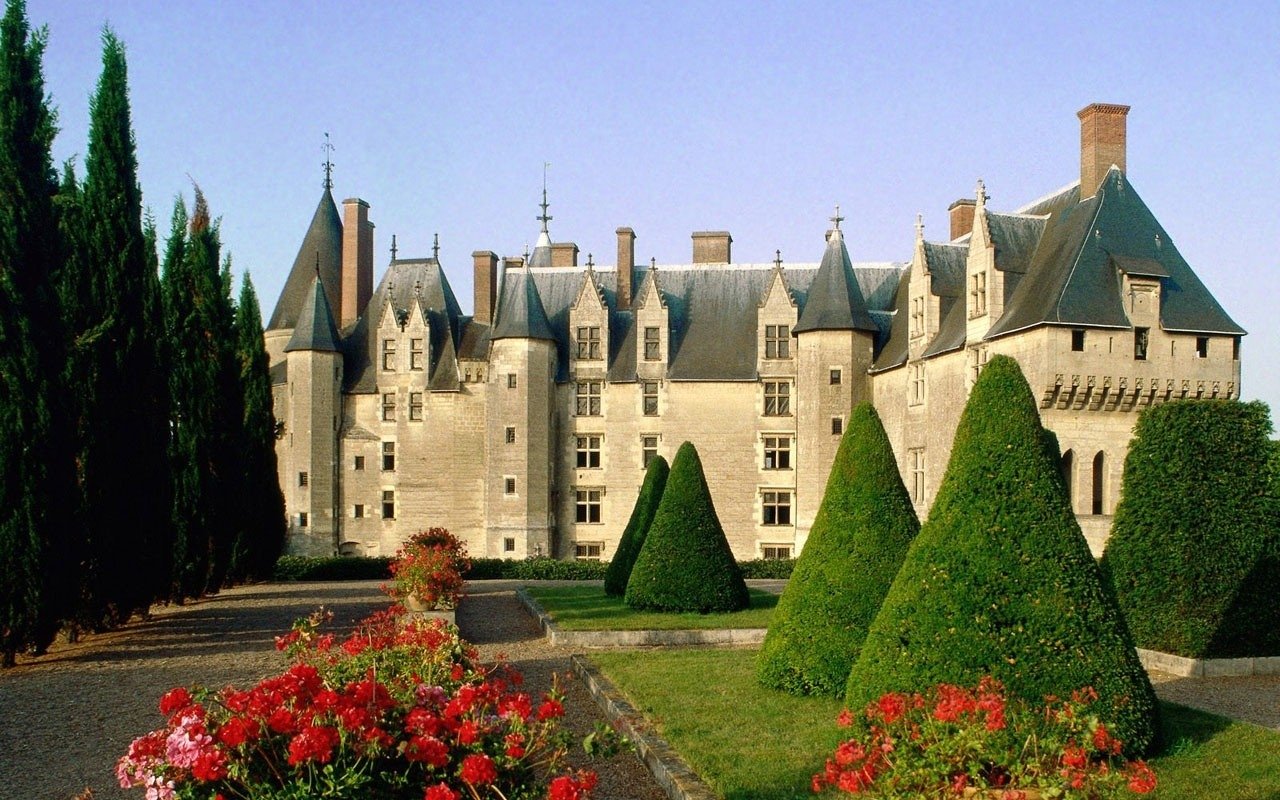 Man-made Château de Langeais: a limestone castle with steep slate roofs and towers, framed by conical topiary and formal flowerbeds under a clear blue sky.