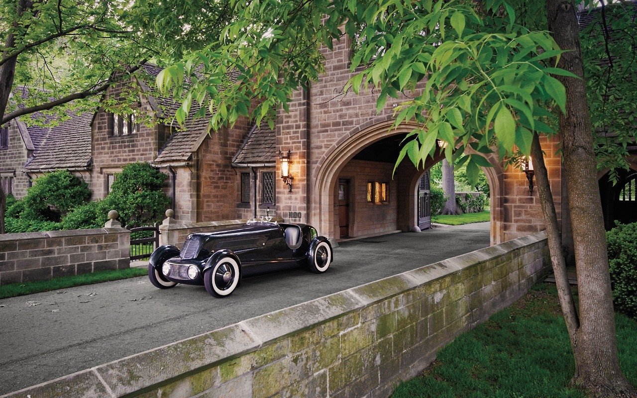 A 1934 Edsel Ford's Model 40 Special Speedster parked on a driveway beside a stone building with arched doorways and surrounding greenery.