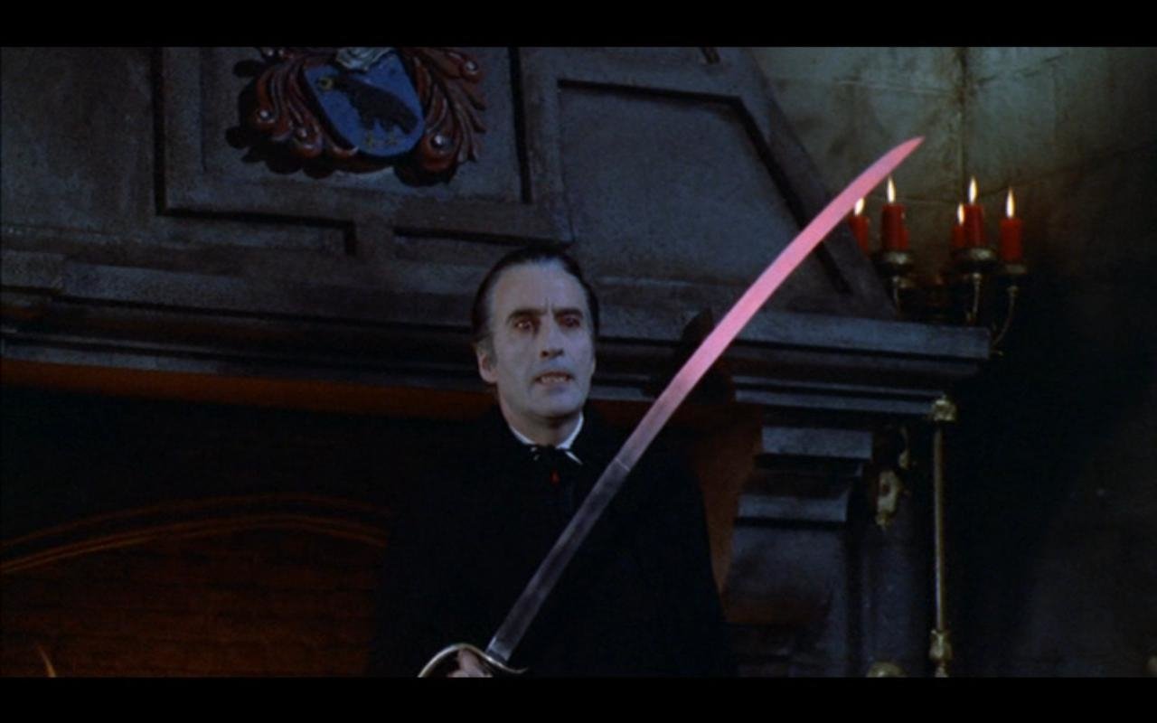 Pale vampire in a dark coat from the movie Scars of Dracula raises a long curved sword in a candlelit Gothic chamber.