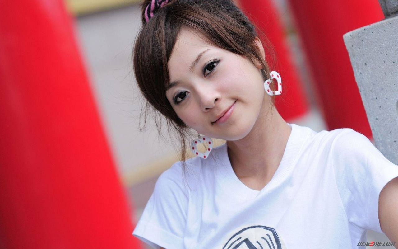 A woman named Mikako Zhang Kaijie poses with a subtle smile, wearing a white shirt and distinctive earrings, set against vibrant red pillars.