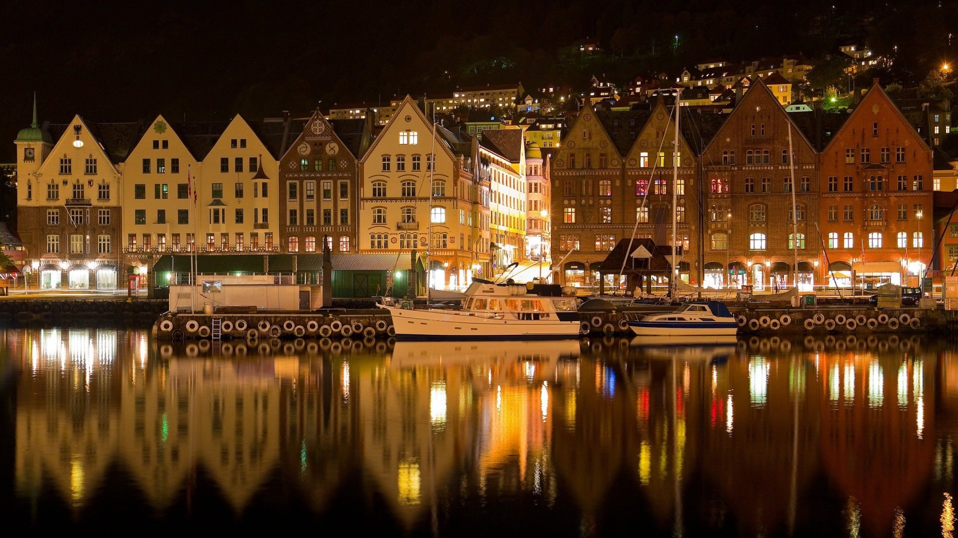 Night view of Bergen's man-made waterfront: row of historic wooden buildings and moored boats glowing with warm lights reflected in calm harbor water.