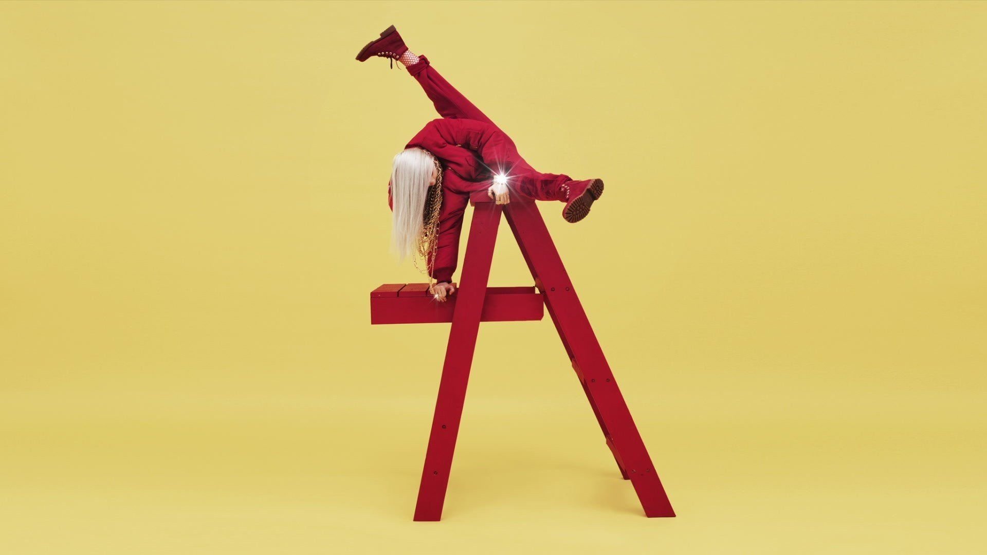 Singer in a red outfit draped dramatically over a red ladder against a yellow backdrop, platinum hair hanging down — bold musical performance pose.