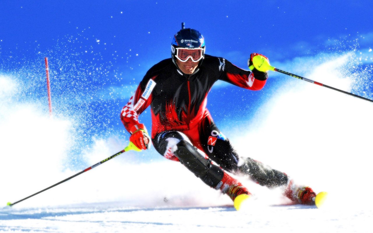 A slalom skier in red and black gear speeds through snowy slopes, skillfully navigating between gates in a dynamic sports action shot.