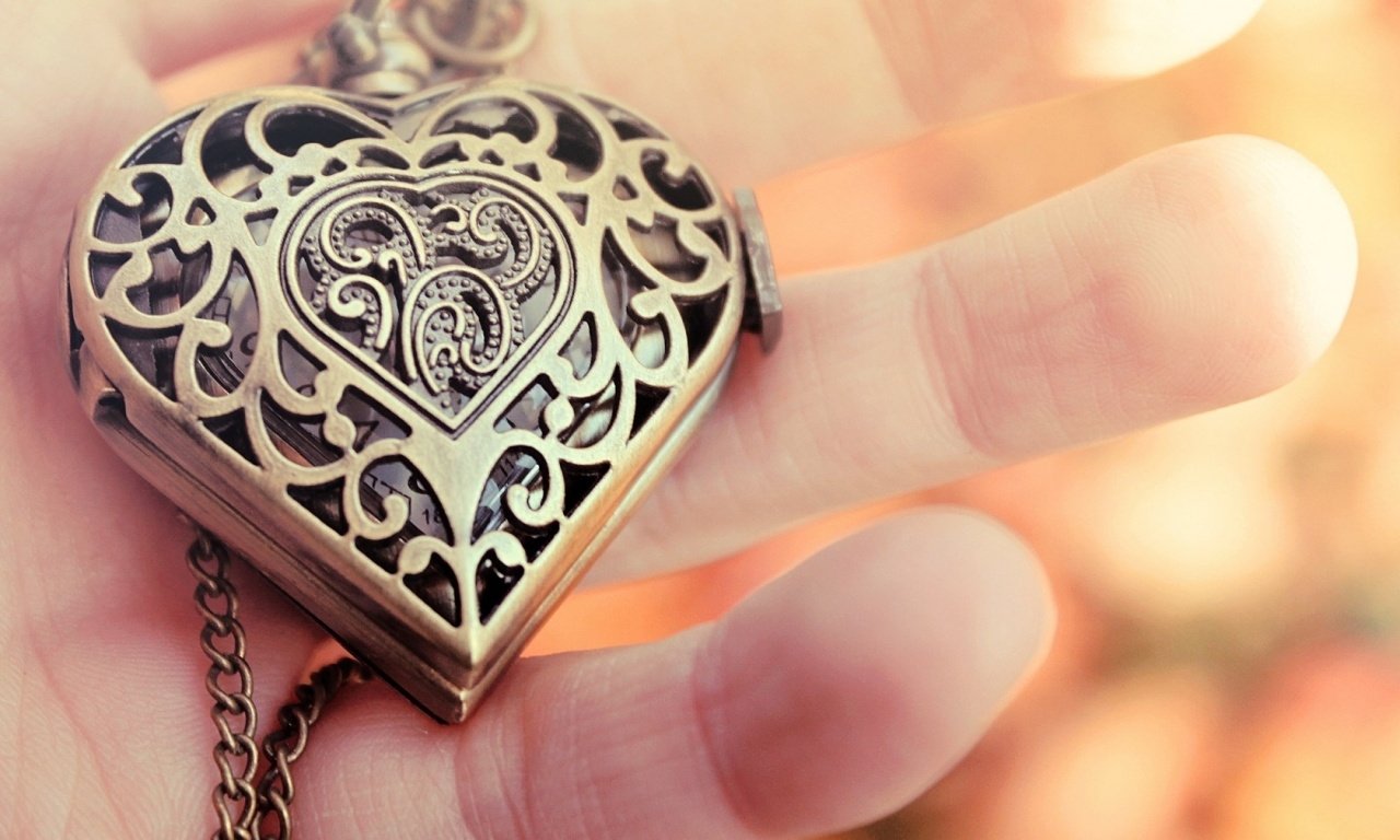 Close-up of man-made jewelry: ornate heart-shaped filigree locket pendant held between fingertips against a warm, blurred background.