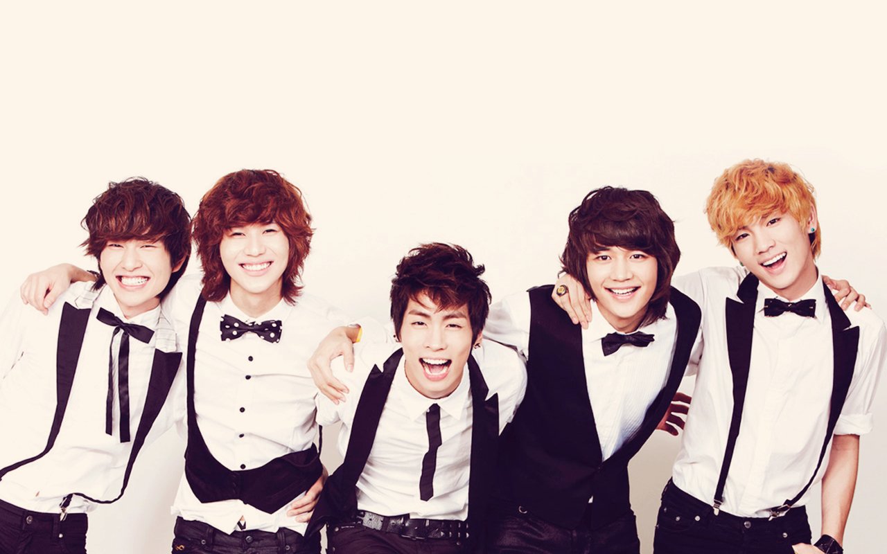 SHINee members dressed in black and white outfits, smiling and posing together against a light background, showcasing their cheerful energy in a music-themed photo.