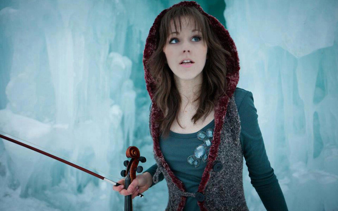Lindsey Stirling playing violin inside an icy cavern, surrounded by blue ice formations.
