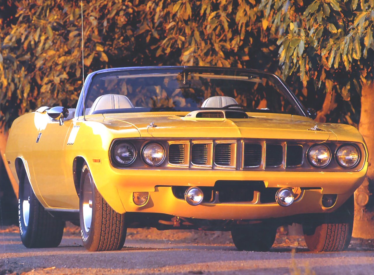 A vibrant yellow Plymouth Hemi Cuda convertible stands on a tree-lined street, showcasing its classic design and powerful presence in the warm sunlight.