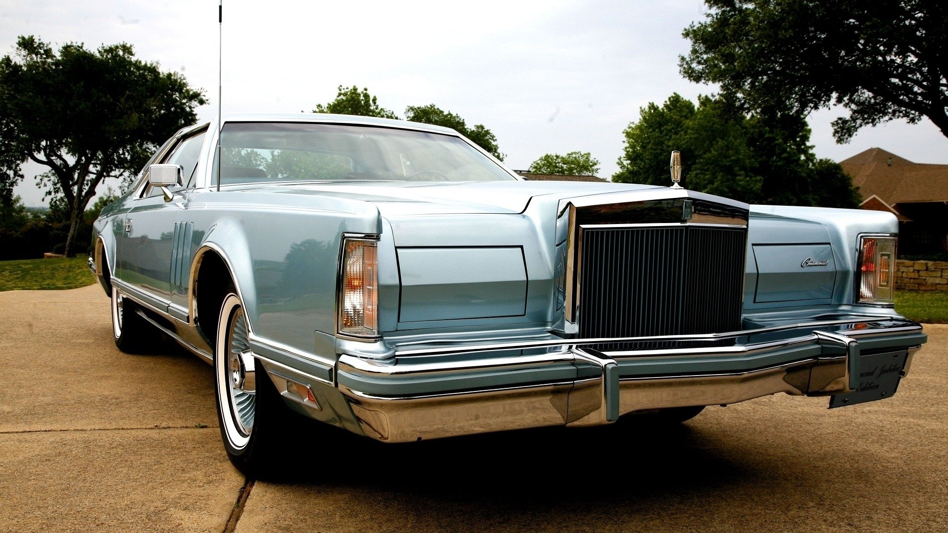 Front three-quarter view of a light blue 1976 Lincoln Continental Mark IV vehicle parked on a driveway, highlighting its chrome grille and whitewall tires.