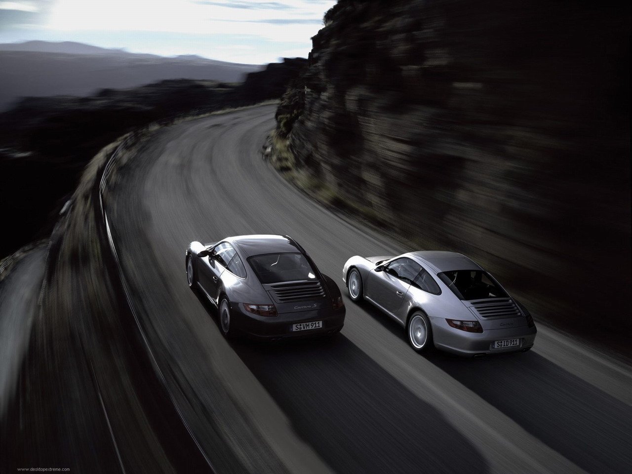 Two Porsche 911 Carrera S vehicles race side by side on a winding mountain road during dusk.