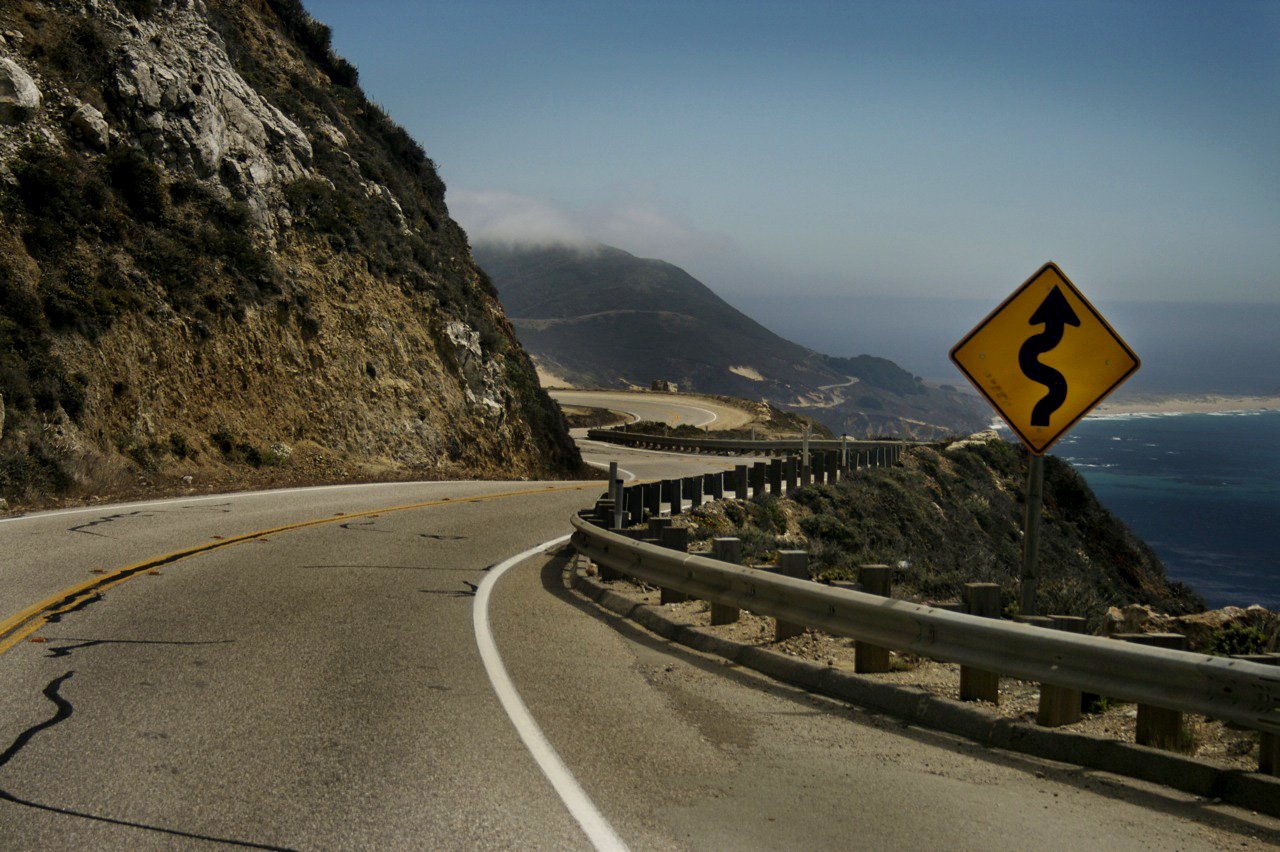Winding man-made road curves along a steep cliffside overlooking the ocean under a clear sky.