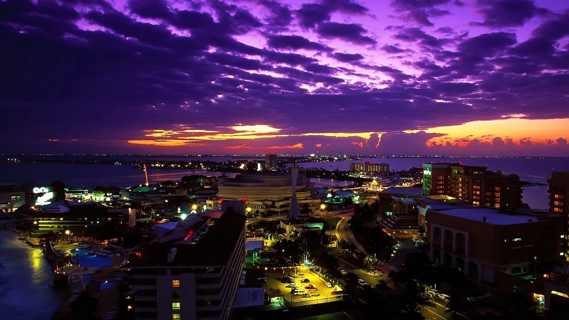 A vibrant purple sunset over the man-made skyline of Cancún, with city lights illuminating buildings and the waterfront.