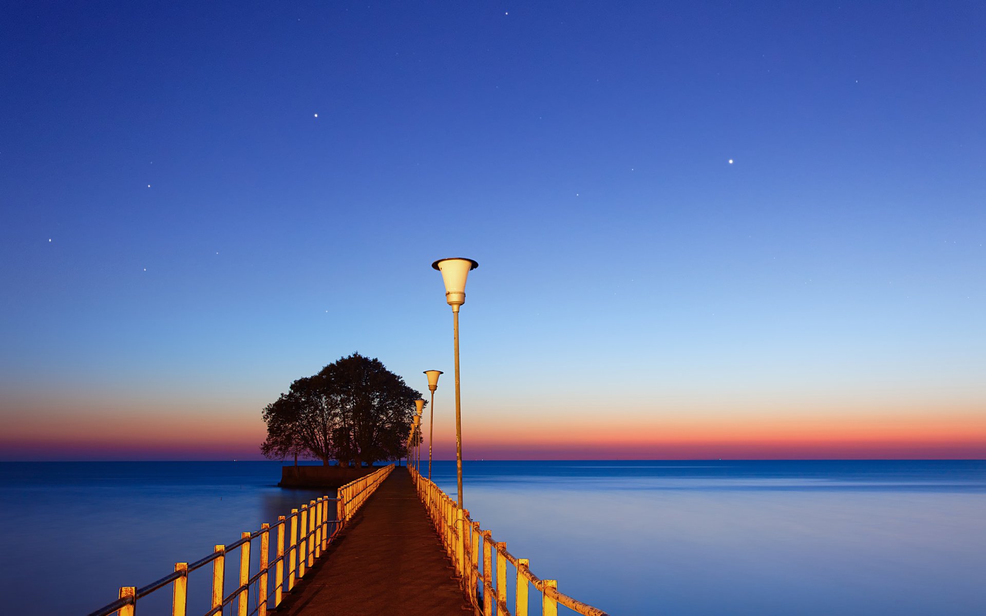 A tranquil man-made pier extends toward a small cluster of trees under a clear twilight sky with street lamps lining the wooden walkway.