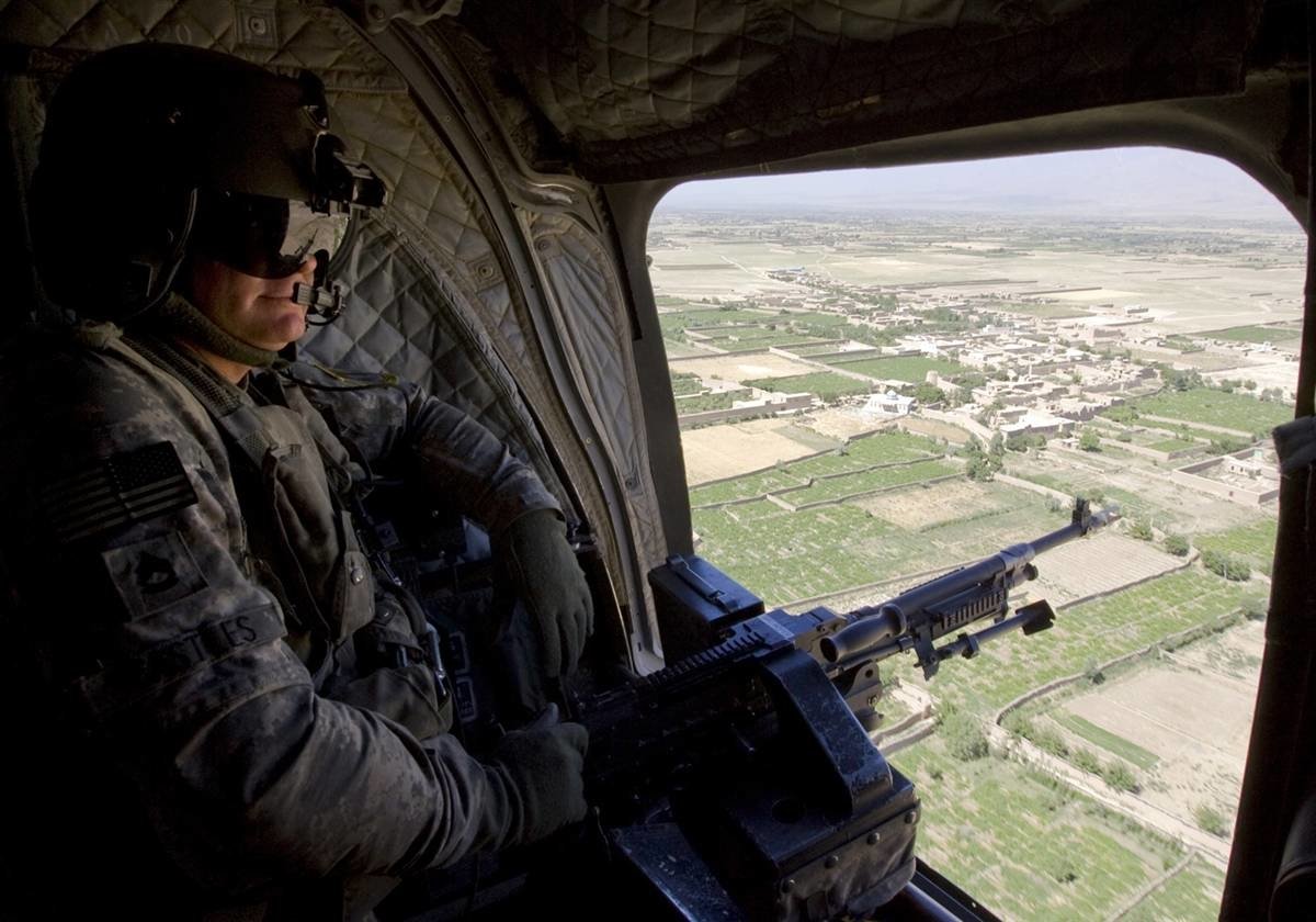 A United States Army soldier sits in a helicopter, equipped with a mounted weapon, overlooking a vast landscape of fields and villages below.