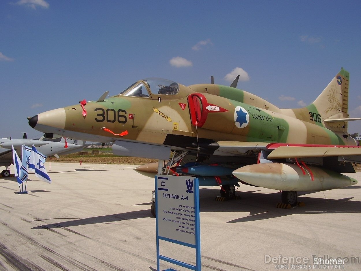 Douglas A-4 Skyhawk military jet painted in desert camouflage, displayed on an airfield with informational signage and Israeli flags nearby.