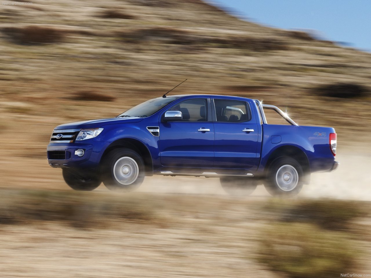 A blue Ford Ranger drives through a rugged landscape, kicking up dust as it navigates the terrain, showcasing its off-road capabilities.
