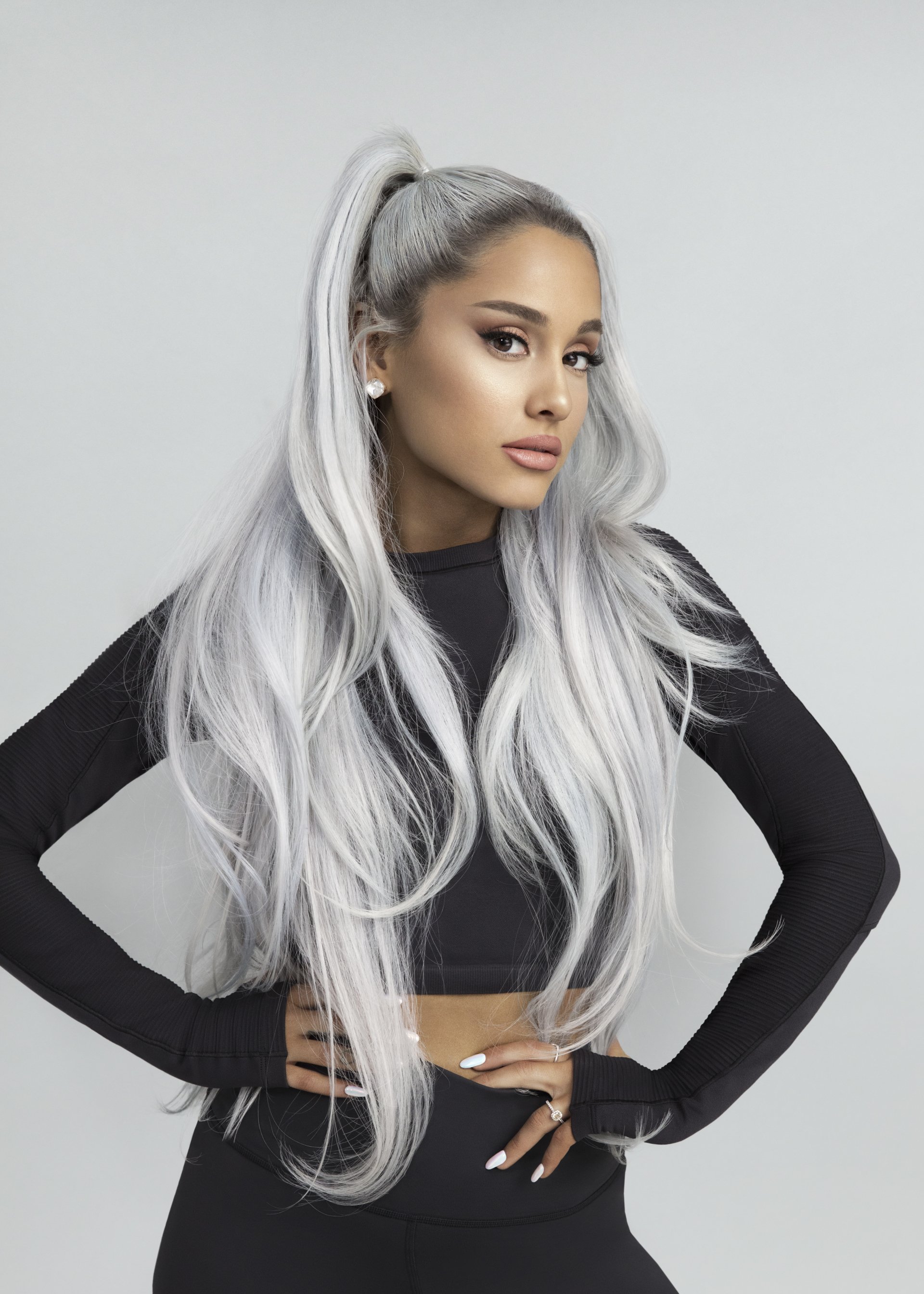 Young woman with long white-silver hair in a high ponytail, wearing a black crop top and leggings, hands on hips against a neutral gray studio background.