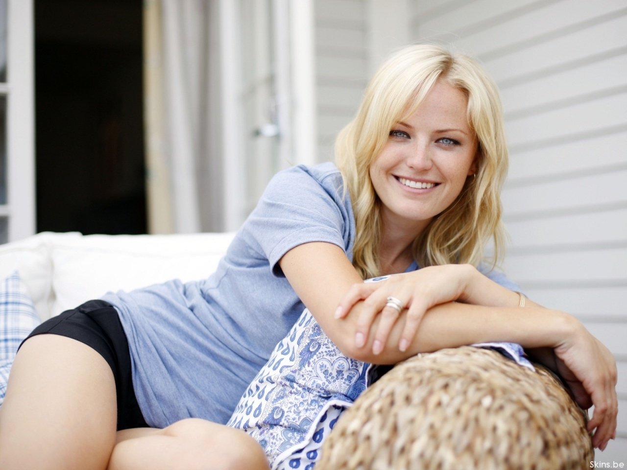 Actress Malin Akerman smiles warmly while reclining on a wicker chair with a patterned cushion on a porch.