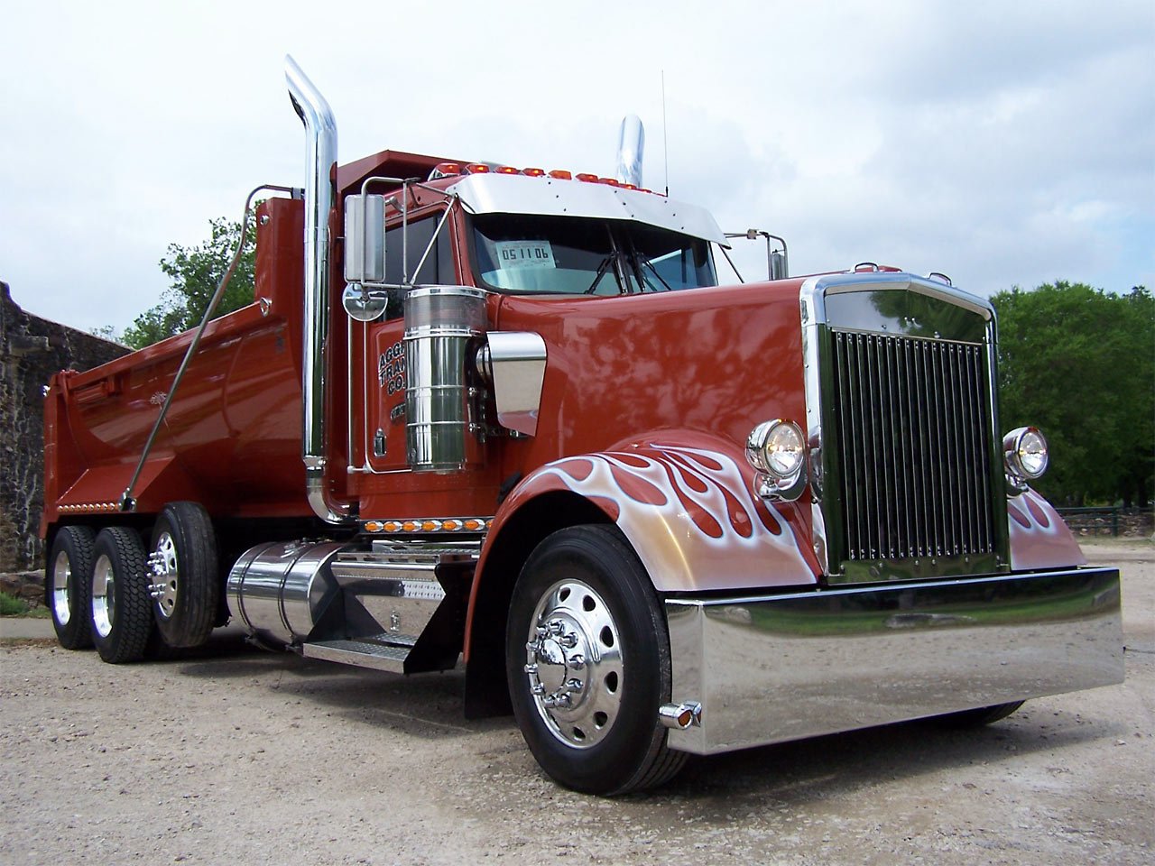 Red Kenworth dump truck with flame decals on the front fenders, parked on a gravel surface with green trees in the background.