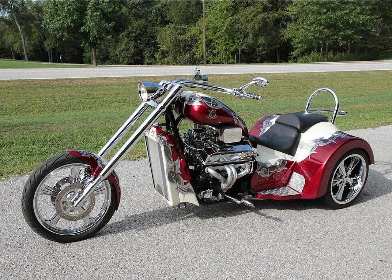 A sleek red and silver trike motorcycle parked on a paved road with a grassy area and trees in the background.