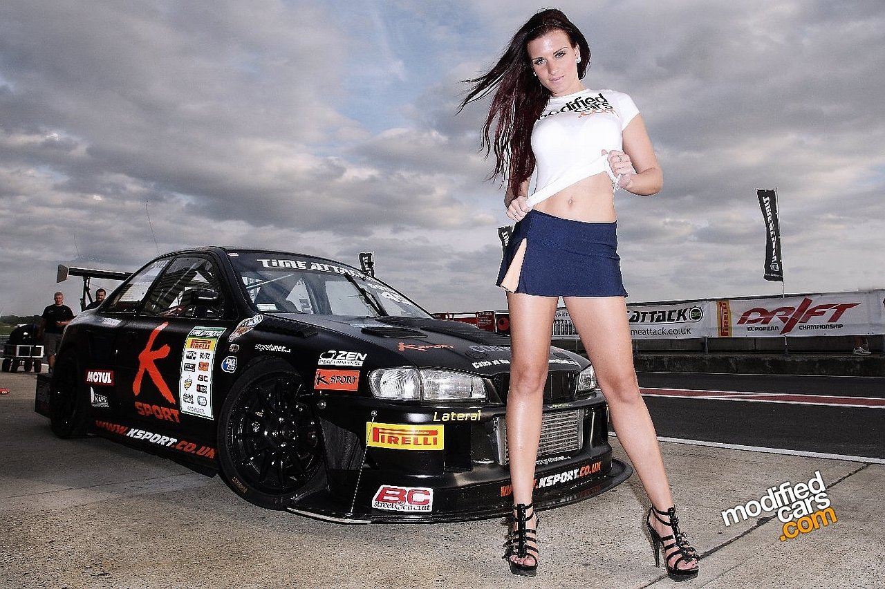 A confident woman stands in front of a modified car, showcasing her style with a cropped top and skirt, embodying the vibrant culture of girls and cars.