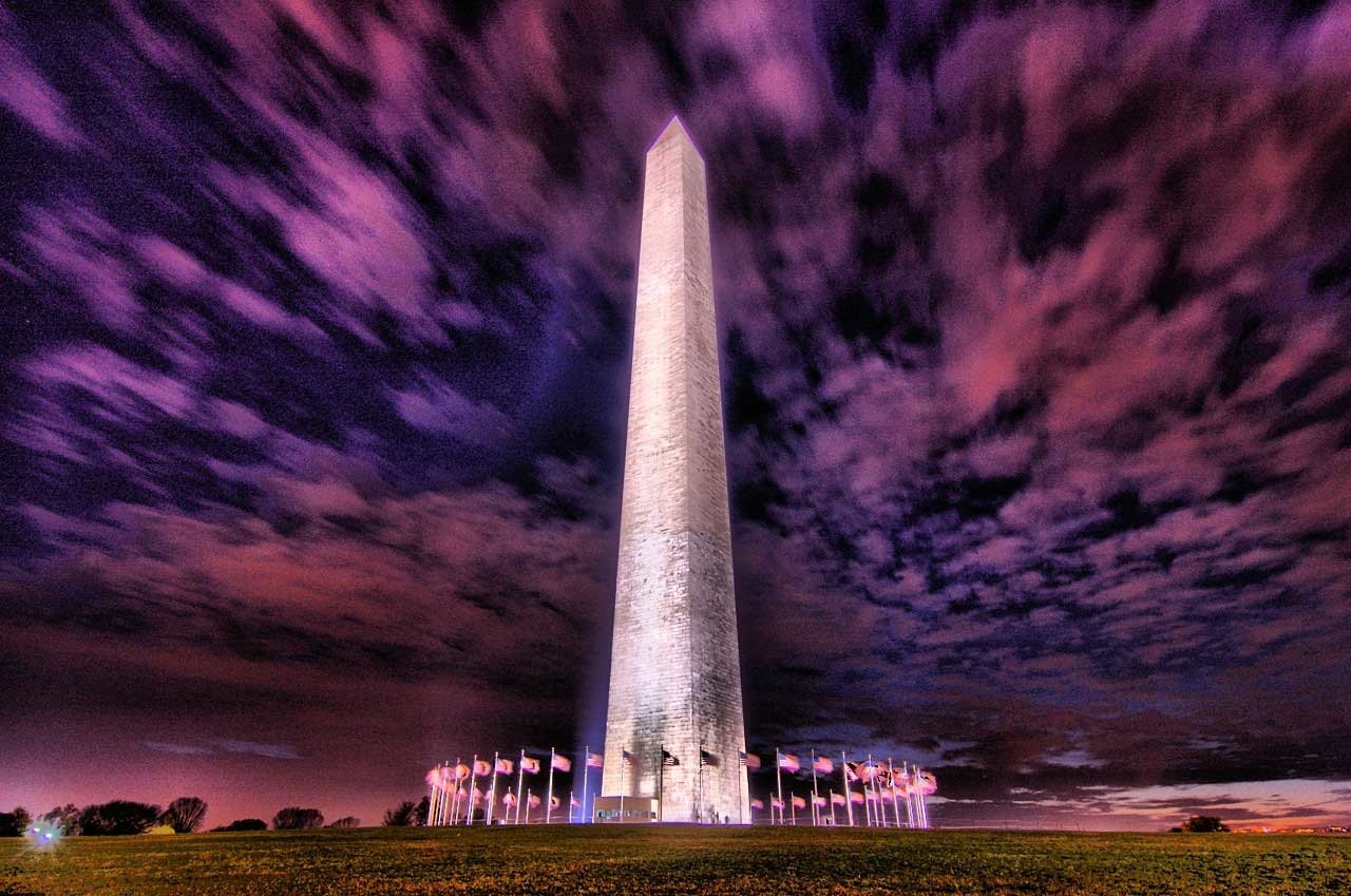 The Washington Monument, a tall man-made obelisk, stands illuminated against a dramatic purple sky with American flags encircling its base.