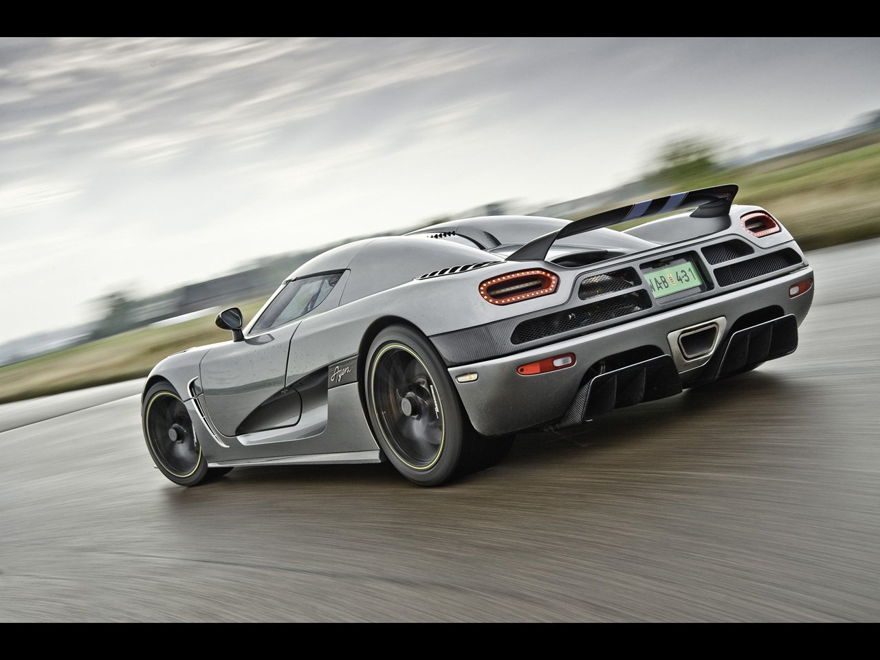 A sleek silver Koenigsegg Agera accelerates on a winding road, showcasing its aerodynamic design and striking rear features against a dynamic backdrop.