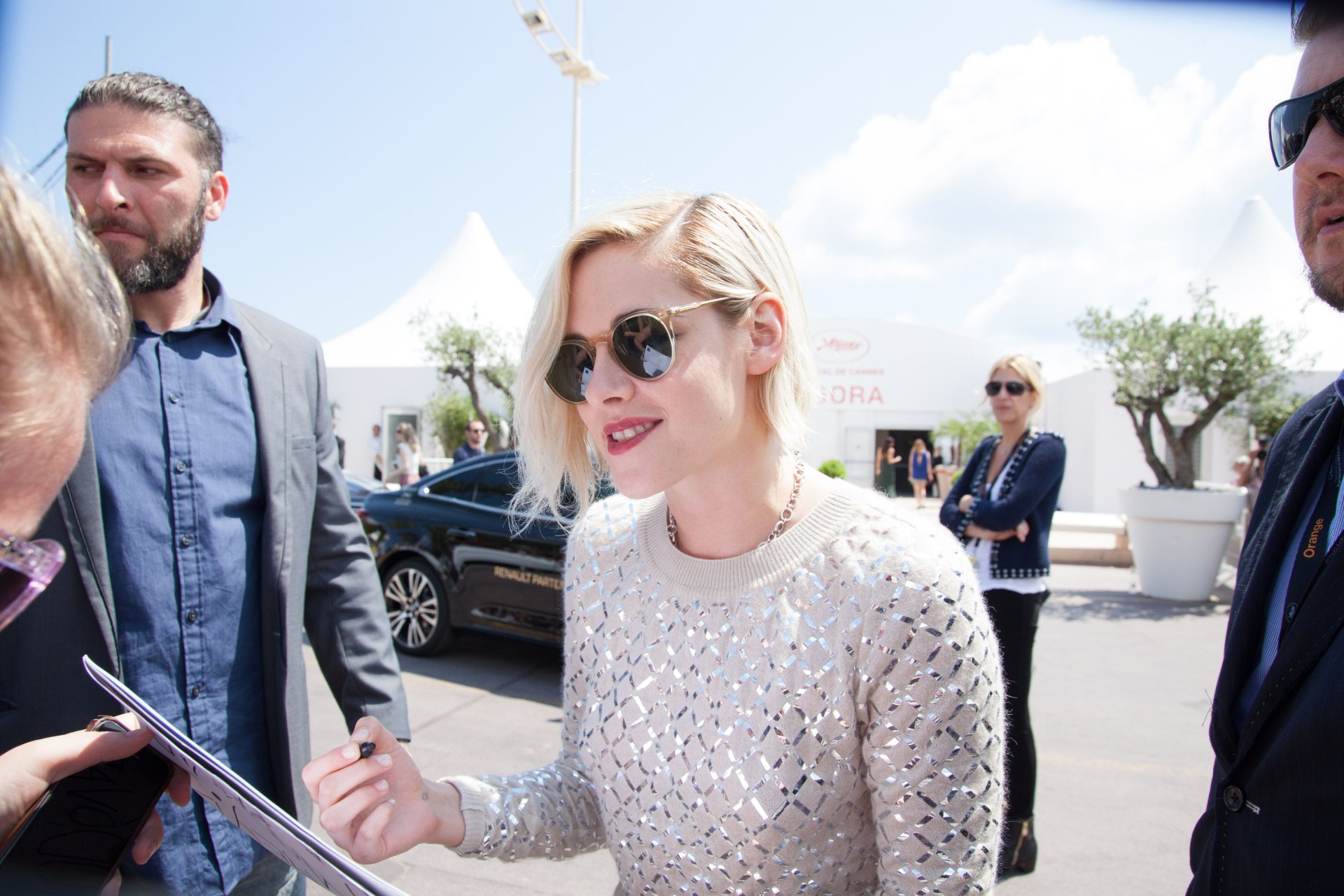 Kristen Stewart, wearing sunglasses and a sparkling top, signs autographs outdoors while surrounded by fans and security on a sunny day.