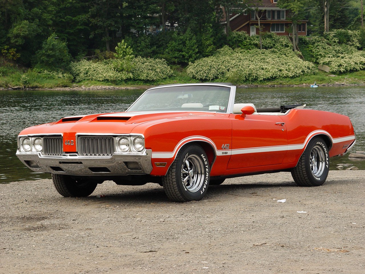 Red Oldsmobile 442 convertible parked near a waterfront with greenery and houses in the background.