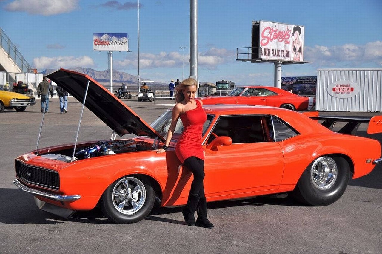 Woman in a red dress poses beside an orange classic muscle car with the hood open at a Girls & Cars event.