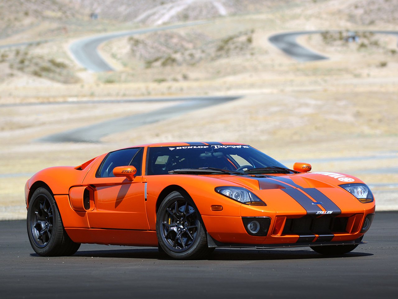 An orange Ford GT sports car with black racing stripes is parked on a winding desert road, showcasing its sleek design against the rugged landscape.