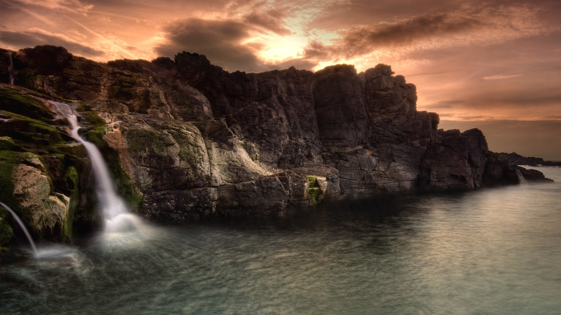 Sunset over rocky coastal cliffs where a slender waterfall cascades into the calm sea — a dramatic nature scene.