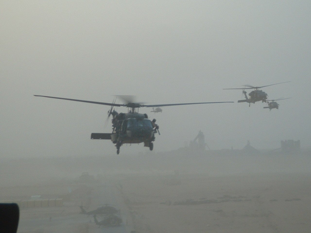 Two Sikorsky UH-60 Black Hawk military helicopters fly low over a dusty landscape with limited visibility.