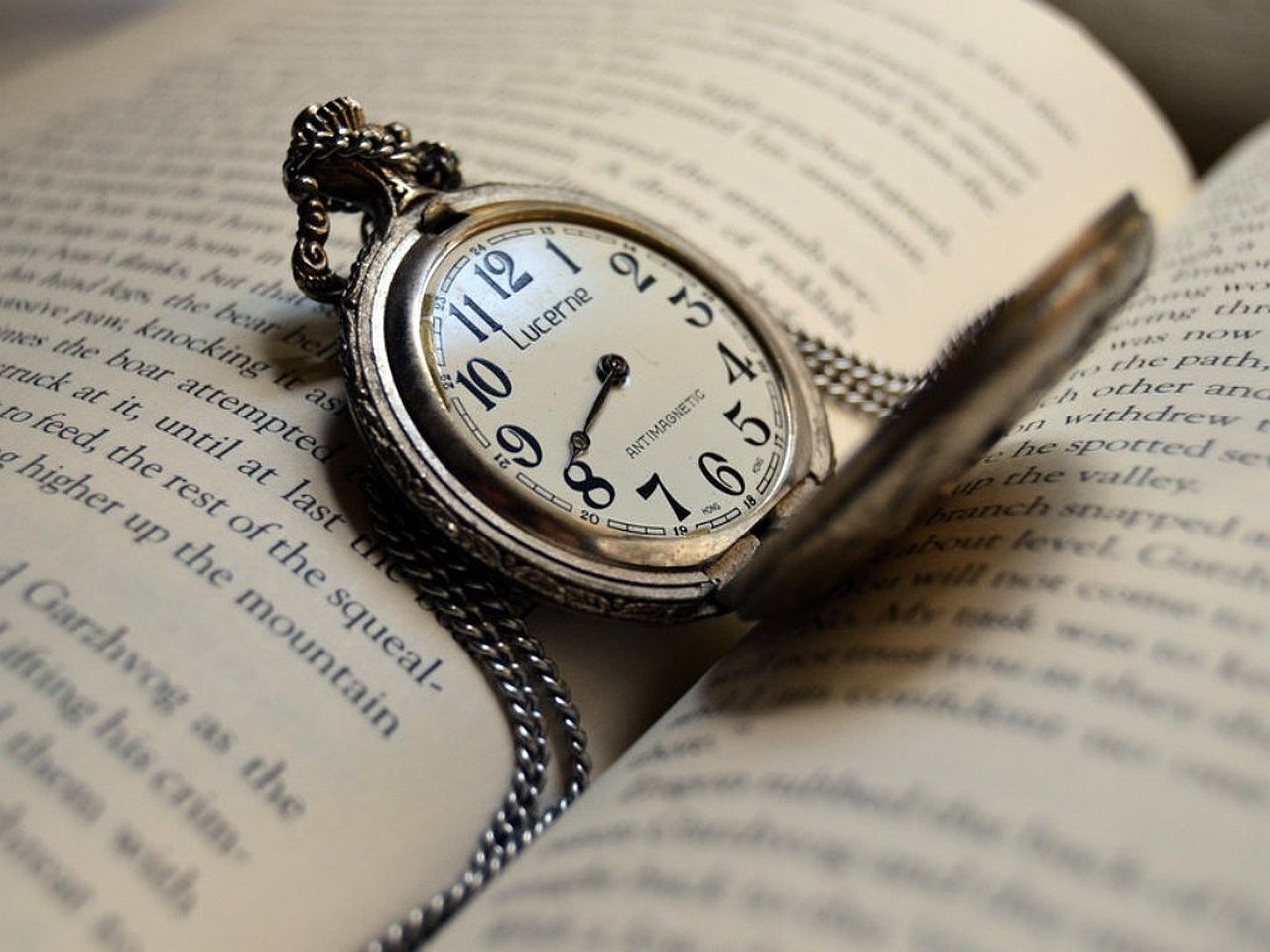 A man-made silver pocket watch with chain and open cover rests on the pages of an open book, its watch face showing about 11:10.